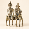 An antique-style metal figurine depicting a seated couple on a woven chair, crafted in a textured metallic finish with warm bronze and silver tones, set against a plain beige background.