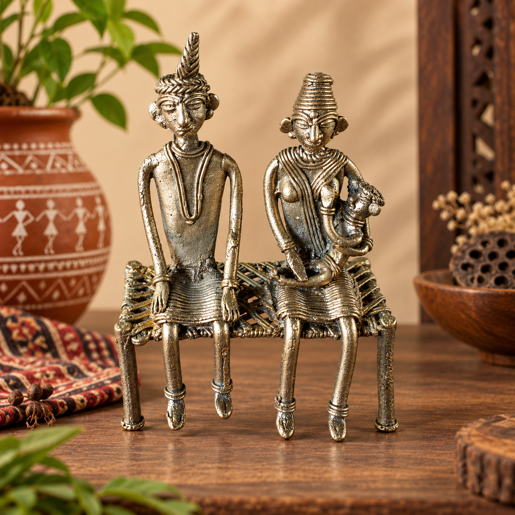 A pair of antique-style metallic brass African tribal figurines depicting a seated man and woman on a woven bench, displayed on a wooden table with decorative background elements.